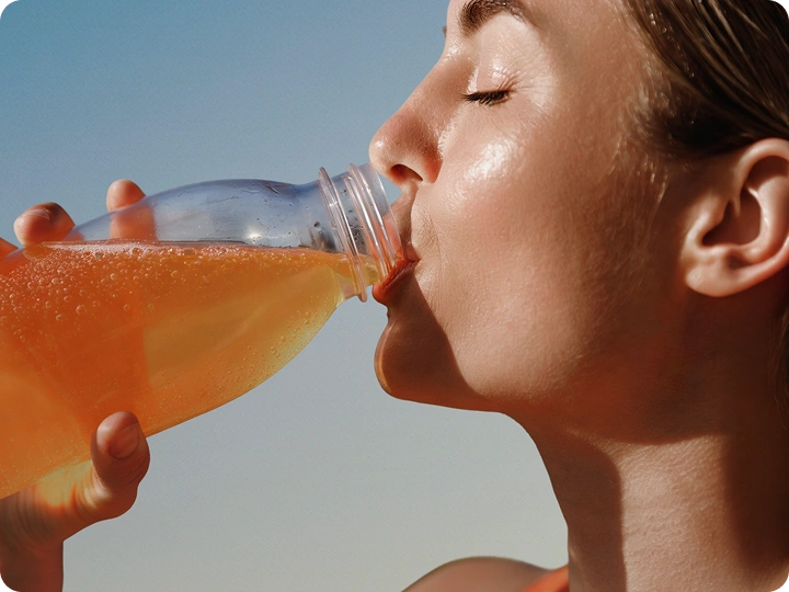 Woman drinking an orange electrolyte beverage from a clear bottle outdoors in sunlight
