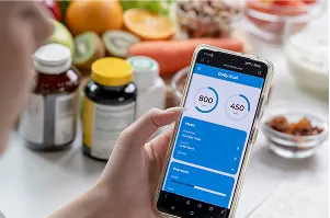 Person using a health tracking app on a smartphone near supplements, fruits, and healthy foods, illustrating wellness tracking, calorie counting, dietary management, and nutritional awareness.