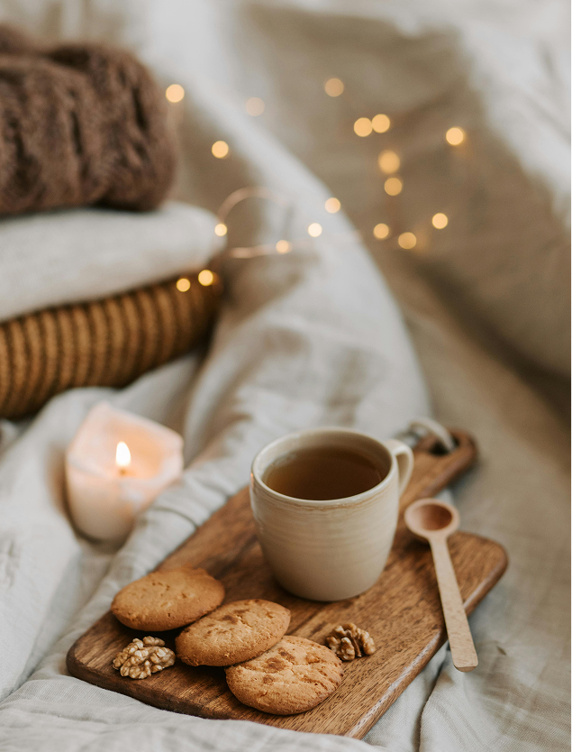 Cozy scene with tea, cookies, walnuts, and candle on tray, surrounded by blankets and warm lights.