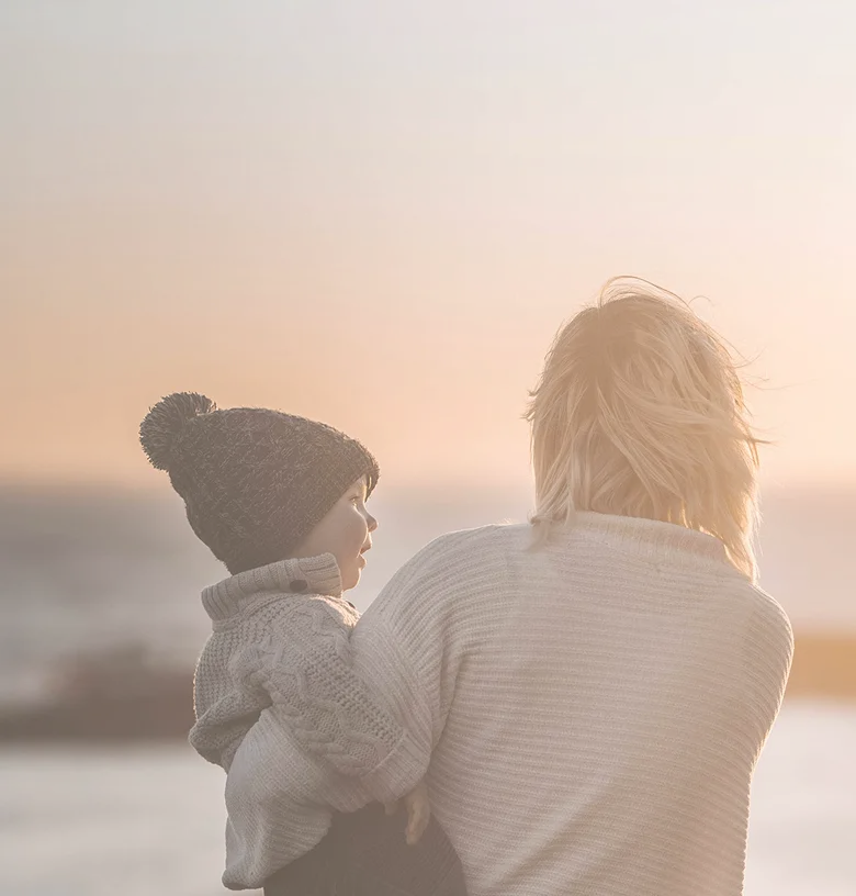 Woman holding child watching sunset over calm sea with warm, glowing sky