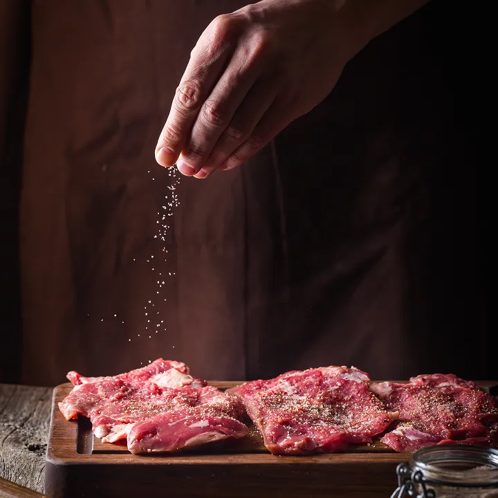 Hand sprinkling seasoning on raw meat on cutting board