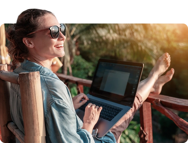 Smiling woman in sunglasses working on a laptop on a sunny outdoor balcony, relaxing with feet up